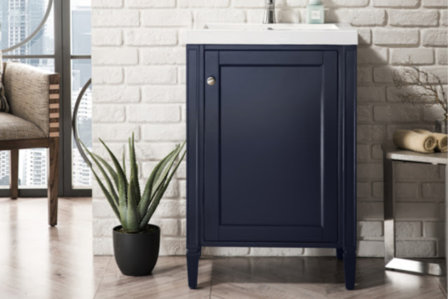 Small single vanity in blue with one cabinet door and a white sink with silver faucet against a white brick wall with chevron patterned wood floor.